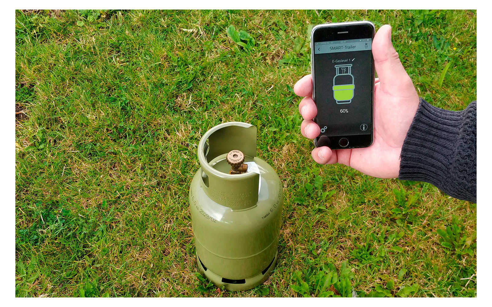 Hand hält Smartphone über einer Gasflasche auf Gras