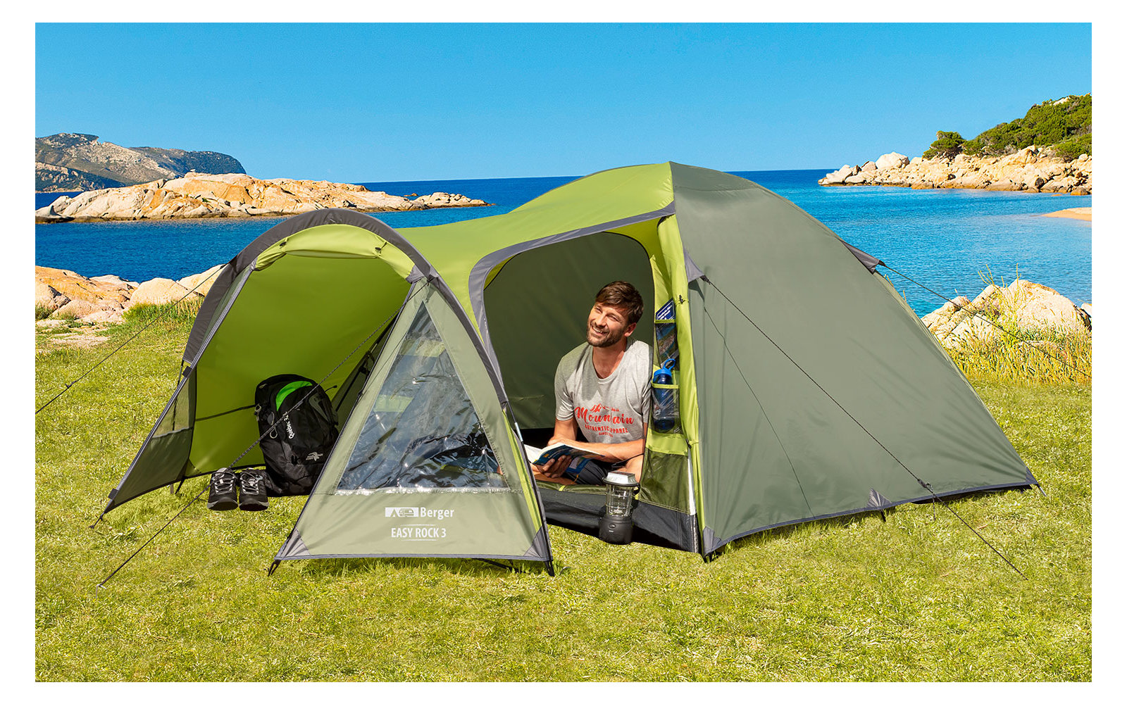 Mann sitzt in einem grünen Zelt auf einer Wiese mit Blick auf einen See und Berge im Hintergrund