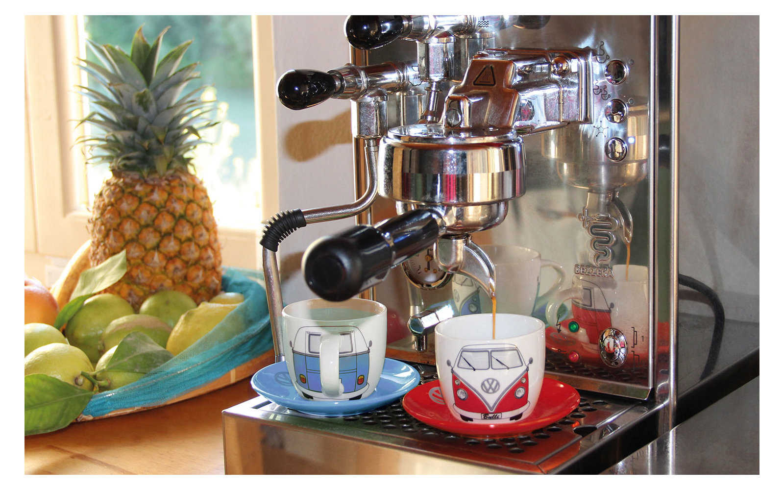 Espressomaschine mit zwei Espressotassen, im Hintergrund Obstschale mit Ananas und Limetten