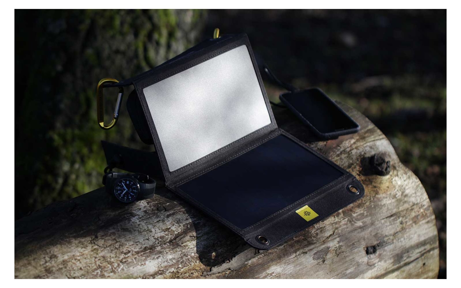 Kleines schwarzes Gerät mit aufklappbarem Bildschirm und gelbem Aufkleber, auf einem Holzstamm liegend, daneben ein Smartphone und ein Objekt