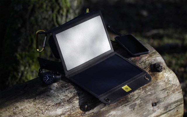 Kleines schwarzes Gerät mit aufklappbarem Bildschirm und gelbem Aufkleber, auf einem Holzstamm liegend, daneben ein Smartphone und ein Objekt