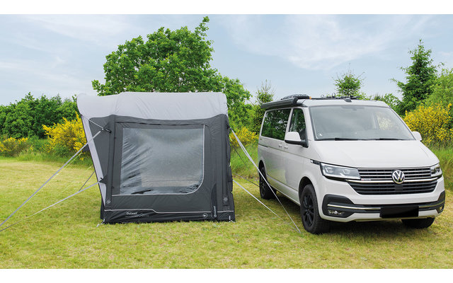 Weißer Campervan neben aufgeschobenem grauen Zelt auf einer grünen Wiese mit Bäumen im Hintergrund