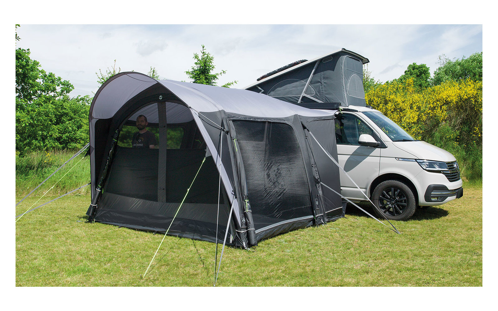 Wohnmobil mit aufgebautem Zelt und Markise auf einer grünen Wiese unter bewölktem Himmel