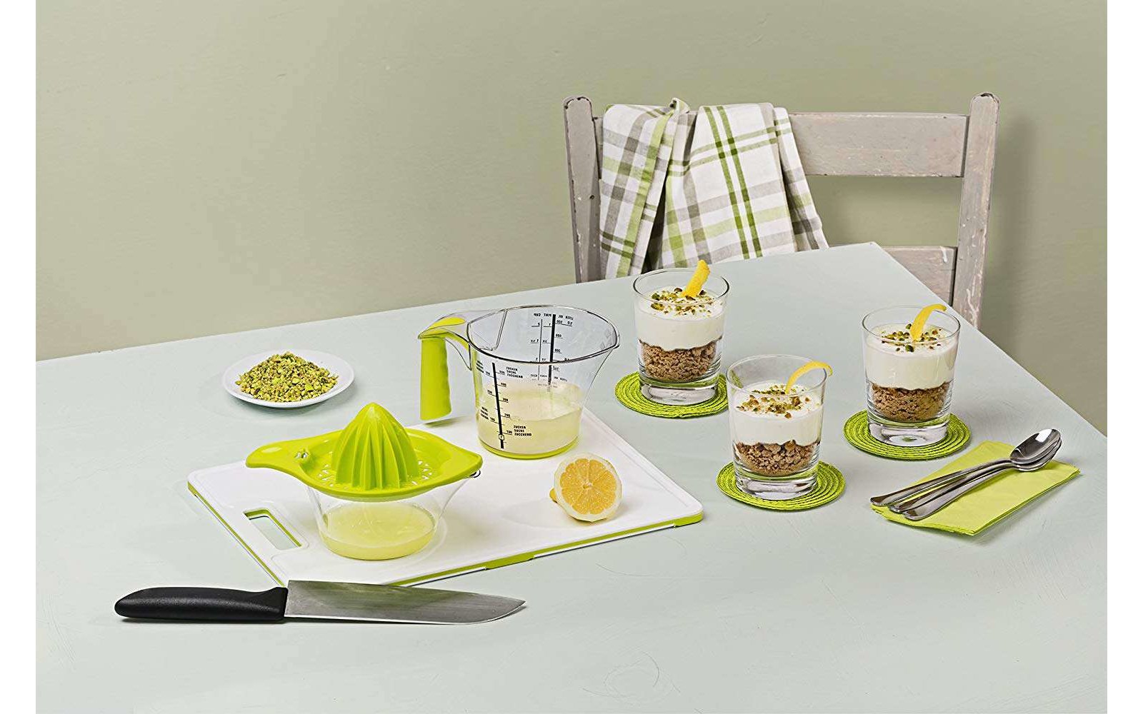 Tisch mit drei kleinen Desserts in Gläsern, Zitronenpresse, Zitrone, Messer und Löffeln, auf einem weißen Tisch mit Stuhl im Hintergrund