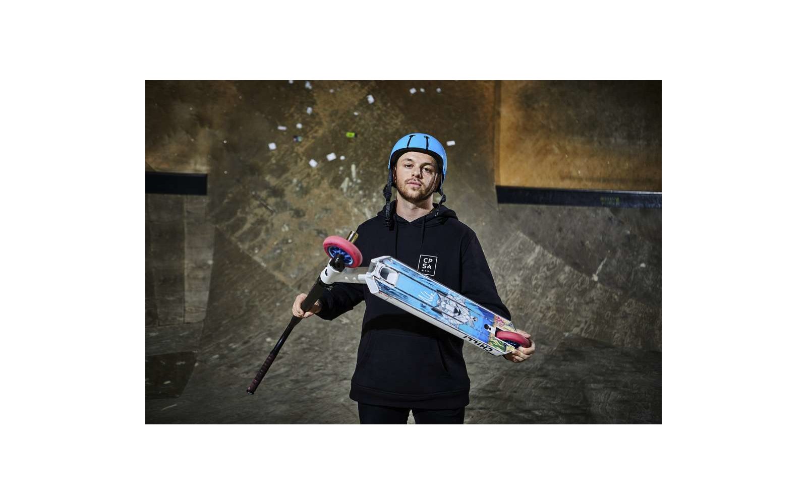 Junger Mann mit Helm hält ein Longboard vor einem Skatepark