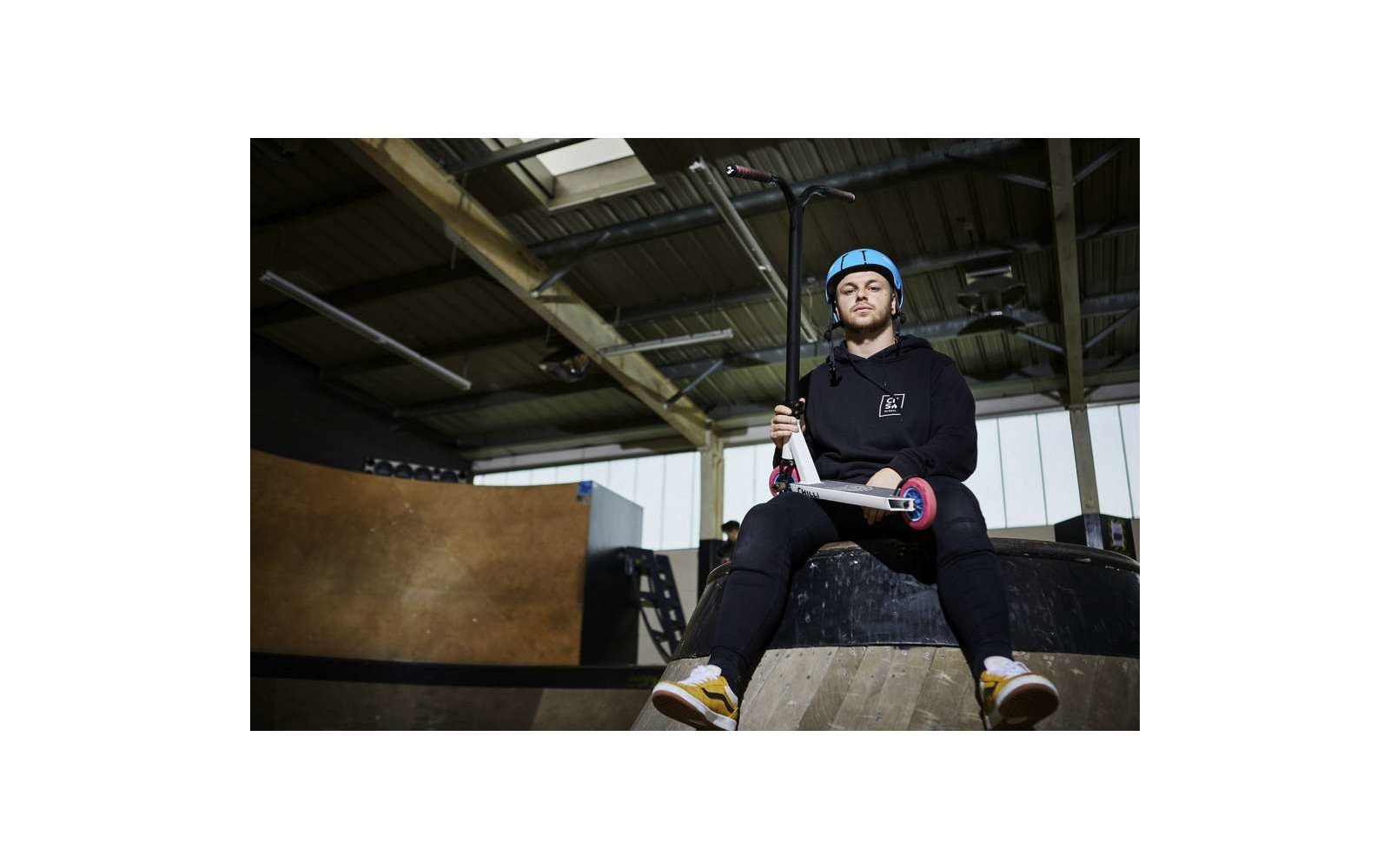 Person mit Helm sitzt auf Rampe in Skatepark und hält ein Skateboard