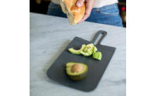 Aufgeschnittene Avocado auf einem schwarzen Schneidebrett, daneben eine Hand, die ein Brötchen hält