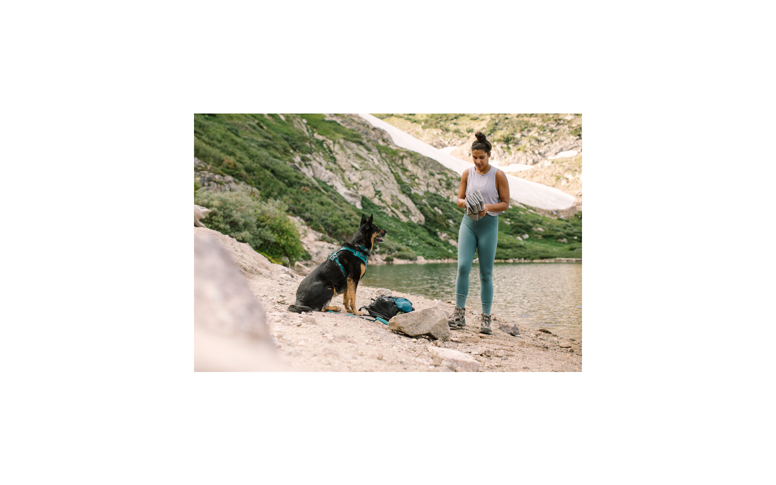 Frau in Sportkleidung steht am Ufer eines Bergsees, ein Hund sitzt auf dem Boden vor ihr