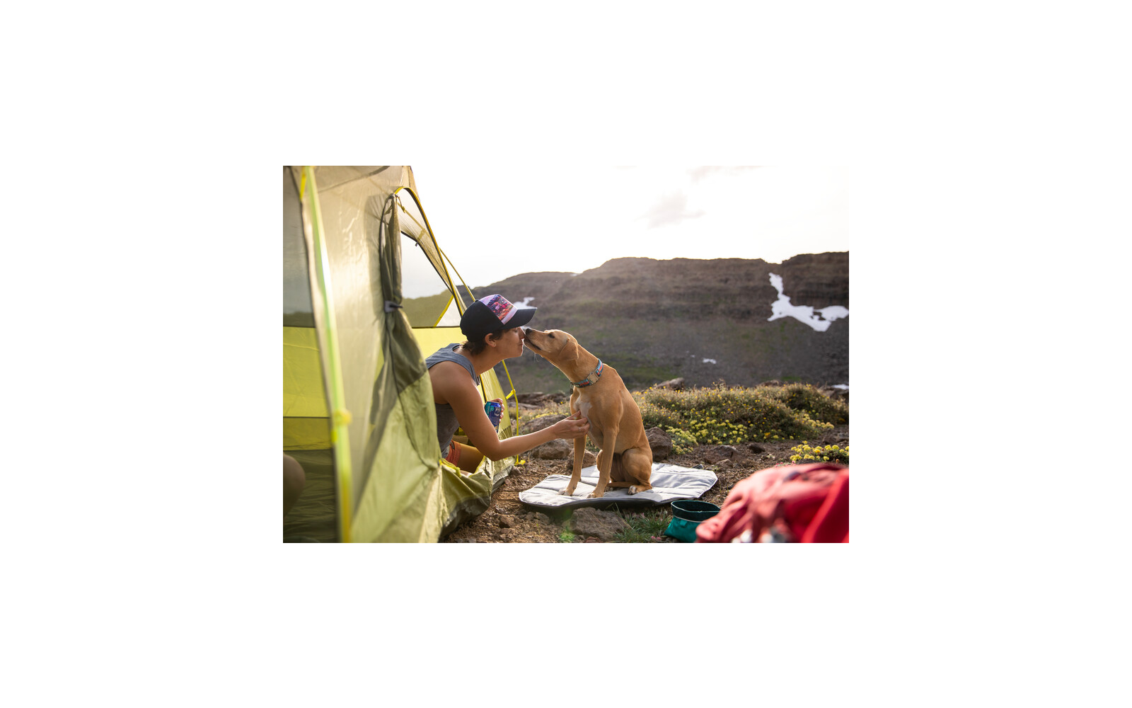 Person in Zelthose kniet und streichelt einen Hund draußen in einer Berglandschaft
