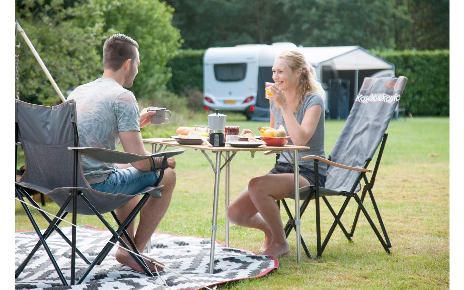 Man and woman sitting at a folding table at a campsite, with a camper van in the background