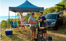 Menschen grillen und sitzen unter einem blauen Pavillon am Wasser, an einem sonnigen Tag