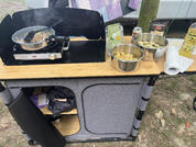 Camping-Küchenzeile auf Gras: tragbarer Gaskocher mit Pfanne, zwei Edelstahlschüsseln mit Nudeln, Papiertuchrolle, Flasche, grüne Verpackung und grauer Aufbewahrungsschrank