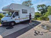 Weißes Wohnmobil mit Offroad-Bike auf einem Anhänger, geparkt an einem sonnigen Parkplatz mit Bäumen im Hintergrund