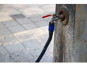 Wasserhahn an einer Metallwand mit angeschlossenem schwarzen Schlauch auf gepflastertem Boden