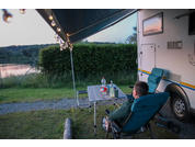Person sitting in a folding chair next to a camper with an awning, overlooking a lake and forested hills in the background
