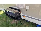Un barbecue noir est posé sur de l'herbe verte à côté d'une caravane blanche