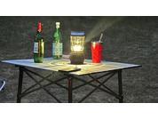 Foldable garden table with two green bottles, a glass, a red can, and brightly lit paper cards at night