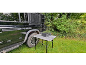 A black vehicle is parked next to a small table on a green lawn with bushes in the background