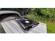 A portable camping stove on a gray wooden table outdoors, with green bushes in the background