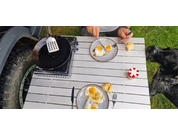 Grill grate, a hand holding tongs, plates with bread rolls, and a red dice game on a garden table