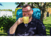 Man met bril zit buiten op een blauwe stoel en drinkt uit een gele mok