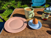 Een houten tafel buiten met een roze bord, een blauwe schotel met een koffiekopje, een ander koffiekopje met patroon en een pot honing