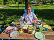 Man zit aan een tuintafel met borden, glazen en verschillende etenswaren in het zonlicht