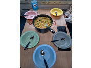 Mesa de madera con una olla de pasta penne y verduras en el centro, cinco platos de colores con cucharas, una botella de agua en el alféizar y parte de una manga a la derecha
