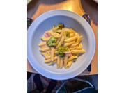 Cuenco blanco con penne, ramilletes de brócoli y pequeños trozos de jamón rosa sobre una mesa de madera, cubiertos en el borde del cuenco