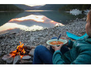 Persona sentada en la orilla pedregosa de un lago de montaña sosteniendo un cuenco de sopa con una cuchara; junto a ella una fogata, un kayak y la montaña reflejada en el agua