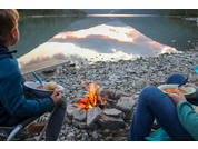 Dos personas sentadas en la orilla pedregosa de un lago; en primer plano una pequeña hoguera; ambas sostienen un cuenco con una cuchara; el lago refleja montañas y el cielo; un muelle de madera en la orilla