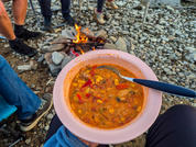 Cuenco rosa con sopa naranja con trozos de verduras y cuchara azul en el regazo de una persona; piernas y zapatos de otras personas alrededor de una pequeña hoguera con llamas sobre terreno pedregoso