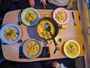 Mesa de madera con cinco cuencos de penne y verduras verdes, una sartén con una cuchara en el centro, cubiertos y manos/antebrazos visibles de personas en la mesa
