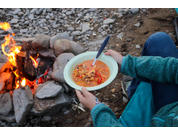 Manos sosteniendo un cuenco de sopa con una cuchara junto a una hoguera sobre terreno rocoso