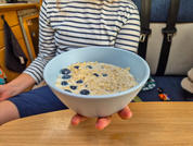 Hand hält hellblaue Schüssel mit Haferbrei und Blaubeeren über einem Holztisch; im Hintergrund sitzt eine Person mit gestreiftem Oberteil