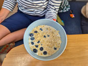 Blaue Schüssel mit Haferbrei und Blaubeeren auf einem Holztisch; teilweise sichtbare sitzende Person mit gestreiftem Shirt und blauen Shorts, Couchkissen und kleines Spielzeug sichtbar
