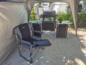 Camping chair and small table in the shade of a tent, grass and trees in the background