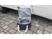 Offene Tasche mit grauem Innenfutter und weißen Tragegriffen auf gepflastertem Boden neben einem weißen Fahrzeug