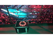 Schwarzer Koffer mit rundem Leuchtring und kleinem Gerät darauf auf einer Tribünenstufe; Metallgeländer im Vordergrund; darunter Zuschauer und hell beleuchtetes Spielfeld mit grüner Beleuchtung