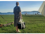 Person mit Dutt steht mit dem Rücken zur Kamera auf Rasen, trägt schwarz-weiß gestreiftes Kleid und hält große schwarz-weiße Tasche; im Hintergrund Meer, Inseln, Zaun, mehrere Personen und ein Sonnenschirm