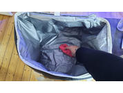 Hand mit schwarzem Ärmel legt roten Lappen in offenen grauen Stoffbehälter auf Holzfußboden; rechts oben eine Wasserflasche sichtbar