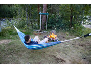 Ein Mann liegt in einer blau-weiß gestreiften Hängematte, trägt dunkles T-Shirt und helle Shorts. Neben ihm brennt ein kleiner Lagerfeuerkreis aus Steinen. Im Hintergrund Bäume, Gras und ein Fluss