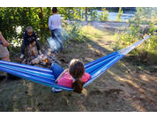 Person liegt in blau-weiß gestreifter Hängematte; zwei weitere Personen stehen und sitzen nahe einem kleinen Lagerfeuer am Ufer eines Sees; Bäume und Rauch im Hintergrund