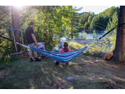 Hängematte mit blau-weißen Streifen zwischen Bäumen; drei Personen: eine links stehend, eine am Boden neben einer kleinen Rauchstelle sitzend, eine in der Hängematte sitzend; See und Bäume im Hintergrund; Holzstapel rechts