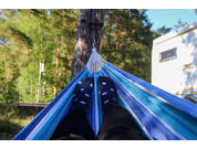 Beine mit schwarzen Socken mit weißen Punkten in einer blau gestreiften Hängematte, Bäume im Hintergrund, weißer Wohnwagen rechts