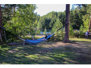 Blaue Hängematte zwischen zwei Bäumen auf einer Wiese, im Hintergrund See und Wald, rechts am Bildrand eine sitzende Person