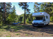 Weißes Wohnmobil rechts auf einer Waldlichtung; links eine blaue Hängematte zwischen Bäumen; Nadelbäume, Gras- und Erdfläche, See im Hintergrund, blauer Himmel