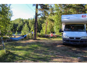 Weißes Wohnmobil rechts, Hängematte zwischen Bäumen links, Personen an einem Picknicktisch, Wald und See im Hintergrund