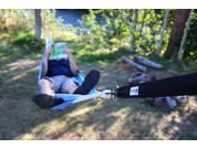 Person liegt in Hängematte und hält ein grünes Buch vor dem Gesicht; Hängemattengurt mit Karabiner im Vordergrund, Gras, Bäume und Wasser im Hintergrund