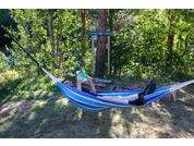 Person liegt in blau-weiß gestreifter Hängematte zwischen Bäumen, hält ein Buch vor das Gesicht; schwarze Schuhe ragen aus der Hängematte; Gras, Holzscheite und ein Baum mit zwei Richtungsschildern im Hintergrund